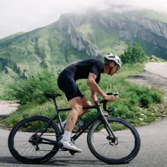 Cycliste en tenue noire gravissant une route sinueuse, profitant de l'ouverture des cols savoyards. Montagnes verdoyantes en arrière-plan.