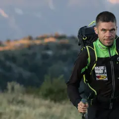 Un homme en tenue de sport, sac à dos et bâtons, progresse sur un trail montagneux au lever du soleil.