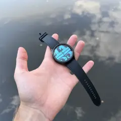 Main d'une personne tenant une Galaxy Watch 4 avec un cadran bleu et des formes géométriques, sur fond d'eau reflétant le ciel. Parfait pour la piscine.