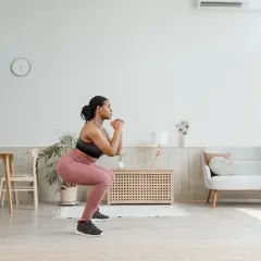 Femme en tenue de sport faisant un squat pour muscler son fessier. Elle est dans un salon lumineux.