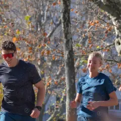 Un homme et une femme souriants courent dans un parc en automne, profitant de leur endurance fondamentale.