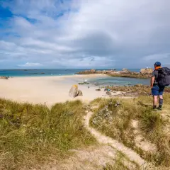 Randonneur contemplant une plage de sable fin, des rochers et la mer. Le sentier GR34 serpente dans les dunes.