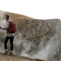 Personne pratiquant la marche nordique, sac à dos rouge, bâtons bleus, contemplant un paysage montagneux brumeux.
