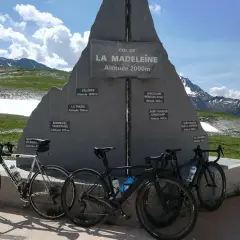 Trois vélos de route devant le monument du col de la Madeleine, une carte des altitudes des villages environnants.