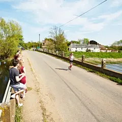 Une femme court sur une route ensoleillée, traversant un pont bordé d'arbres. Des spectateurs encouragent la course à pied en Bretagne.