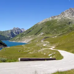 Route sinueuse menant au col du Glandon, avec un lac bleu vif et des montagnes verdoyantes sous un ciel clair.