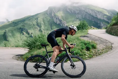 Cycliste en tenue noire gravissant une route sinueuse, profitant de l'ouverture des cols savoyards. Montagnes verdoyantes en arrière-plan.