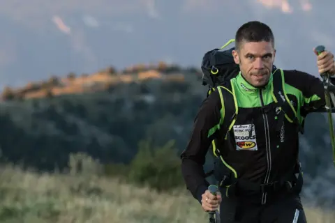 Un homme en tenue de sport, sac à dos et bâtons, progresse sur un trail montagneux au lever du soleil.