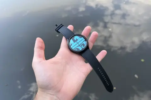 Main d'une personne tenant une Galaxy Watch 4 avec un cadran bleu et des formes géométriques, sur fond d'eau reflétant le ciel. Parfait pour la piscine.