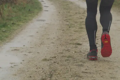 Jambes d'un coureur sur un chemin de terre, concentré sur sa cadence course à pied. Les chaussures rouges et noires s'enfoncent dans le sol humide.