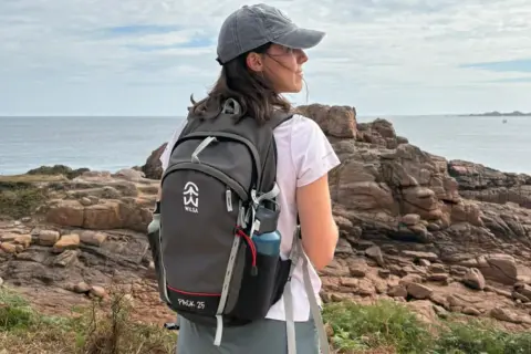 Jeune femme avec un sac à dos, prête à faire de la randonnée sur un sentier côtier rocheux, face à la mer.