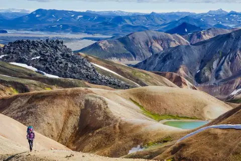 Randonneuse solitaire explorant les paysages colorés et volcaniques du trek Landmannalaugar, avec des montagnes aux teintes ocre, vertes et bleues.