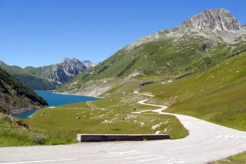 Route sinueuse menant au col du Glandon, avec un lac bleu vif et des montagnes verdoyantes sous un ciel clair.