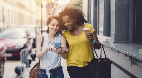 Deux amies marchent en ville, l'une montre son téléphone, l'autre mange une glace. Elles atteignent sûrement leurs 10000 pas par jour !