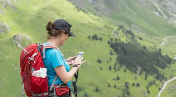 Femme avec sac à dos rouge et bâtons de marche, consultant son téléphone pour sa randonnée 1 journée dans les montagnes verdoyantes.