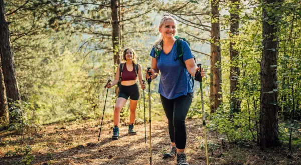 Deux femmes souriantes randonnent en forêt avec des bâtons, profitant des marche avec bâton bienfaits pour la santé et le bien-être.