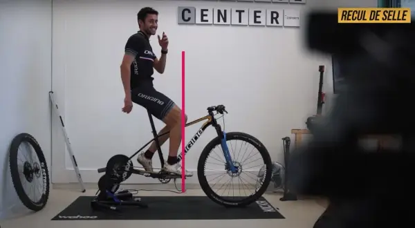 Un cycliste ajuste le recul de selle de son vélo sur un home trainer. Une ligne rose indique la mesure.