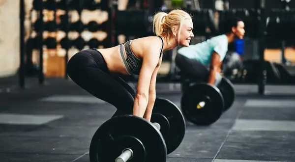 Femme blonde souriante en tenue de sport, prête à soulever une barre. Un homme s'entraîne en arrière-plan. Un entraînement de cardio intense.