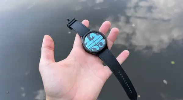 Main d'une personne tenant une Galaxy Watch 4 avec un cadran bleu et des formes géométriques, sur fond d'eau reflétant le ciel. Parfait pour la piscine.