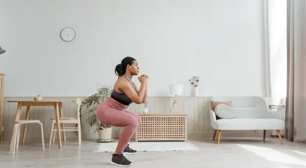 Femme en tenue de sport faisant un squat pour muscler son fessier. Elle est dans un salon lumineux.