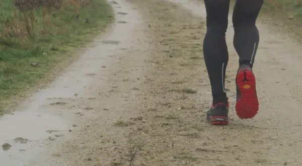 Jambes d'un coureur sur un chemin de terre, concentré sur sa cadence course à pied. Les chaussures rouges et noires s'enfoncent dans le sol humide.