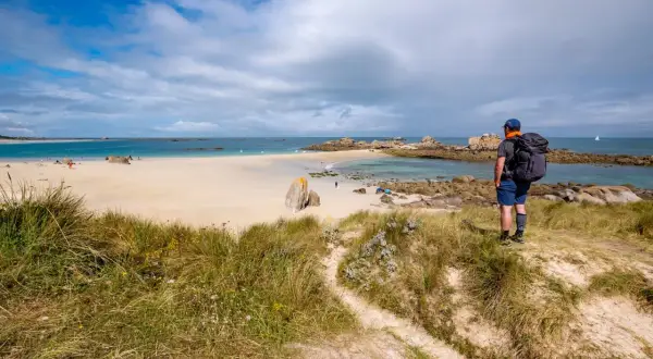Randonneur contemplant une plage de sable fin, des rochers et la mer. Le sentier GR34 serpente dans les dunes.