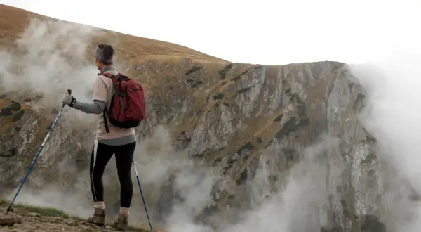Personne pratiquant la marche nordique, sac à dos rouge, bâtons bleus, contemplant un paysage montagneux brumeux.