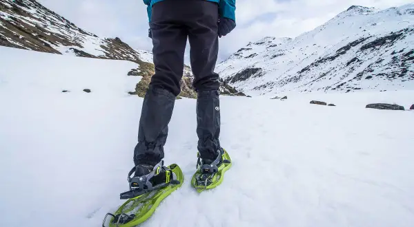 Une personne en tenue d'hiver est prête à marcher avec des raquettes dans un paysage enneigé et montagneux.