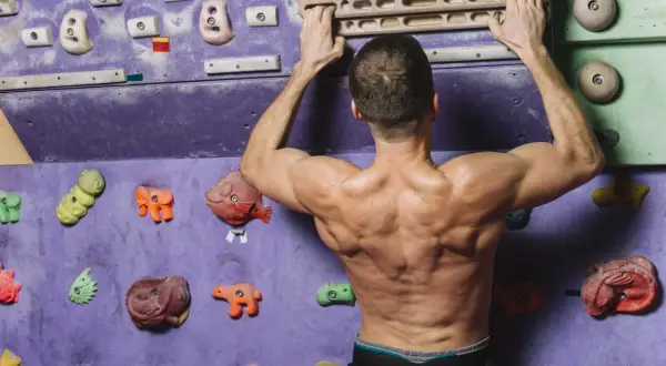 Un grimpeur musclé s'entraîne sur un mur d'escalade coloré, saisissant une barre en bois pour un entraînement intensif.