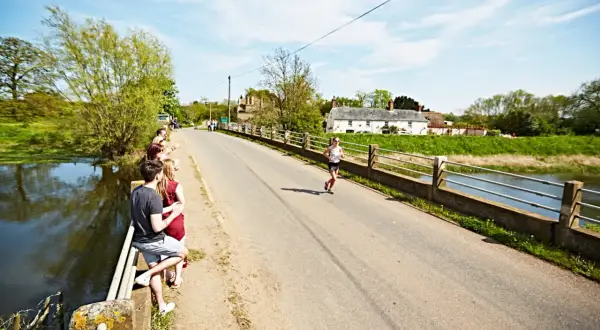 Une femme court sur une route ensoleillée, traversant un pont bordé d'arbres. Des spectateurs encouragent la course à pied en Bretagne.