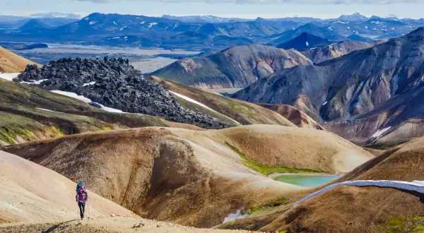 Randonneuse solitaire explorant les paysages colorés et volcaniques du trek Landmannalaugar, avec des montagnes aux teintes ocre, vertes et bleues.