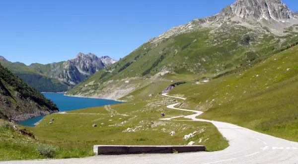Route sinueuse menant au col du Glandon, avec un lac bleu vif et des montagnes verdoyantes sous un ciel clair.