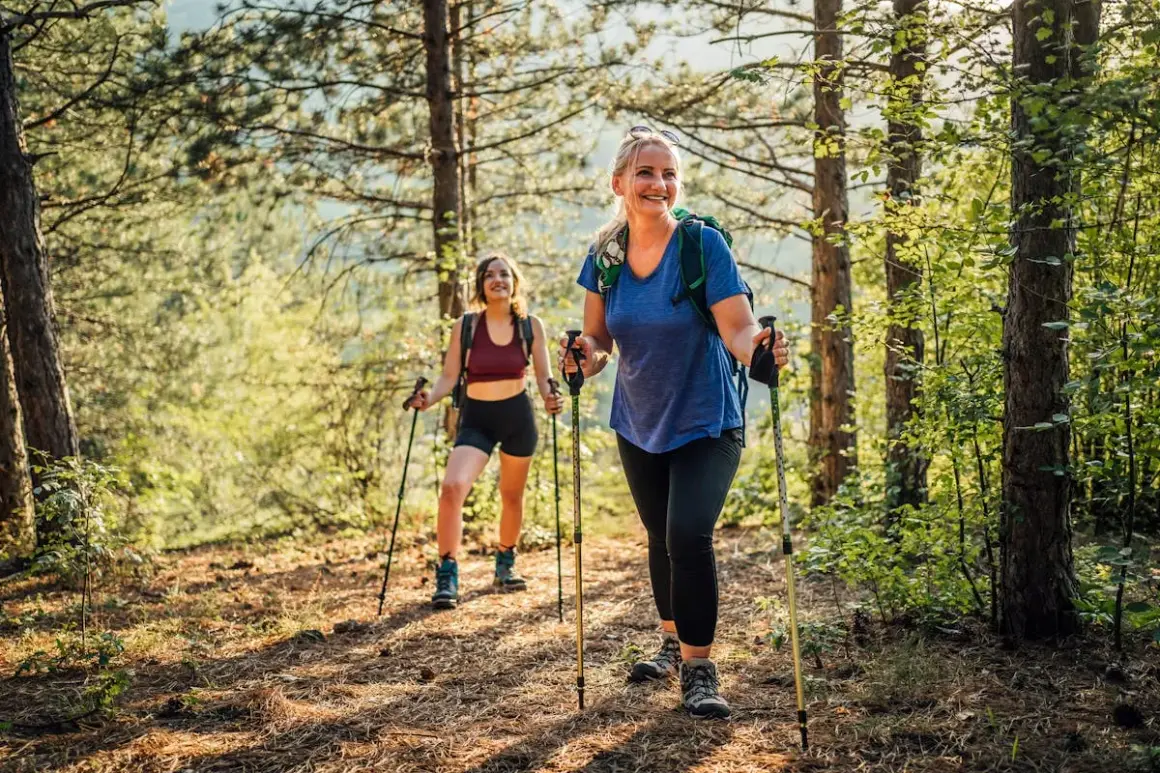 Deux femmes souriantes randonnent en forêt avec des bâtons, profitant des marche avec bâton bienfaits pour la santé et le bien-être.