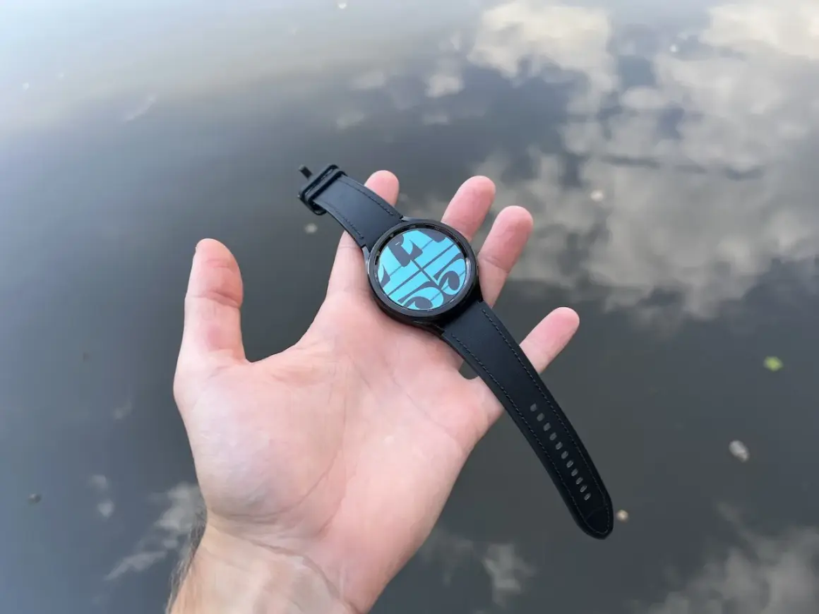 Main d'une personne tenant une Galaxy Watch 4 avec un cadran bleu et des formes géométriques, sur fond d'eau reflétant le ciel. Parfait pour la piscine.