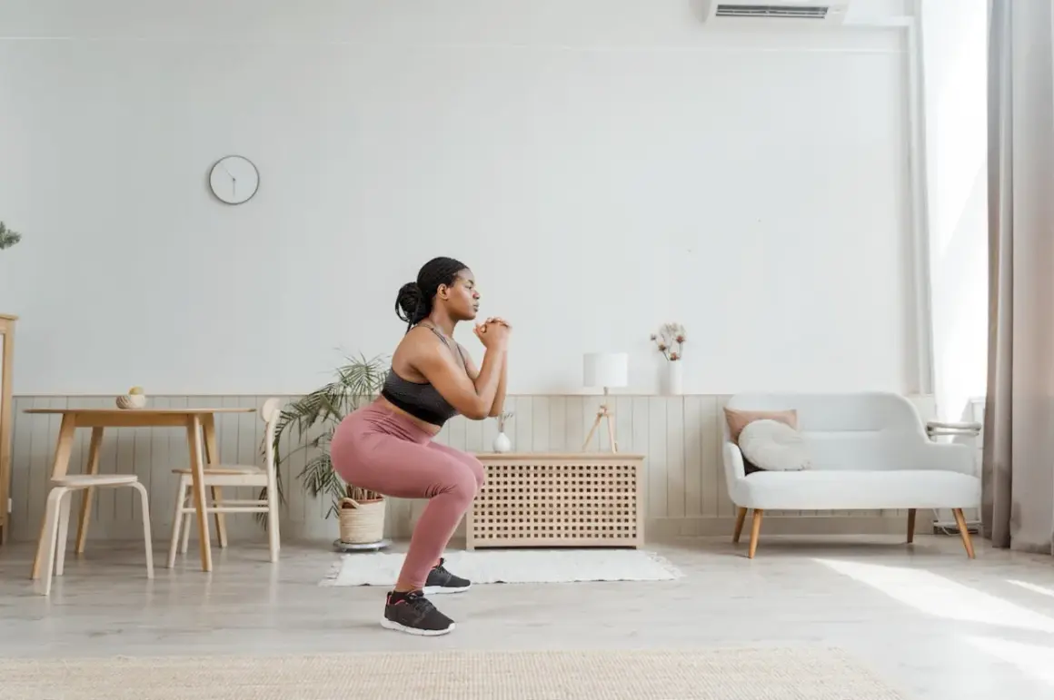 Femme en tenue de sport faisant un squat pour muscler son fessier. Elle est dans un salon lumineux.