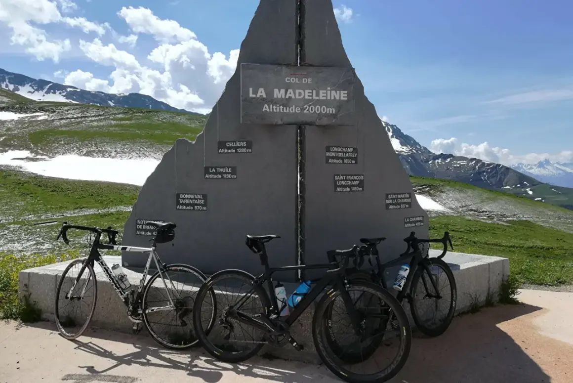 Trois vélos de route devant le monument du col de la Madeleine, une carte des altitudes des villages environnants.