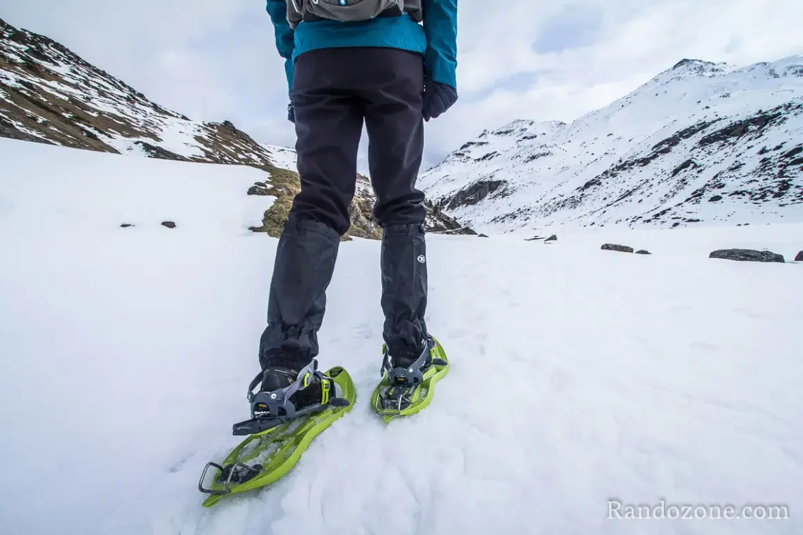Une personne en tenue d'hiver est prête à marcher avec des raquettes dans un paysage enneigé et montagneux.