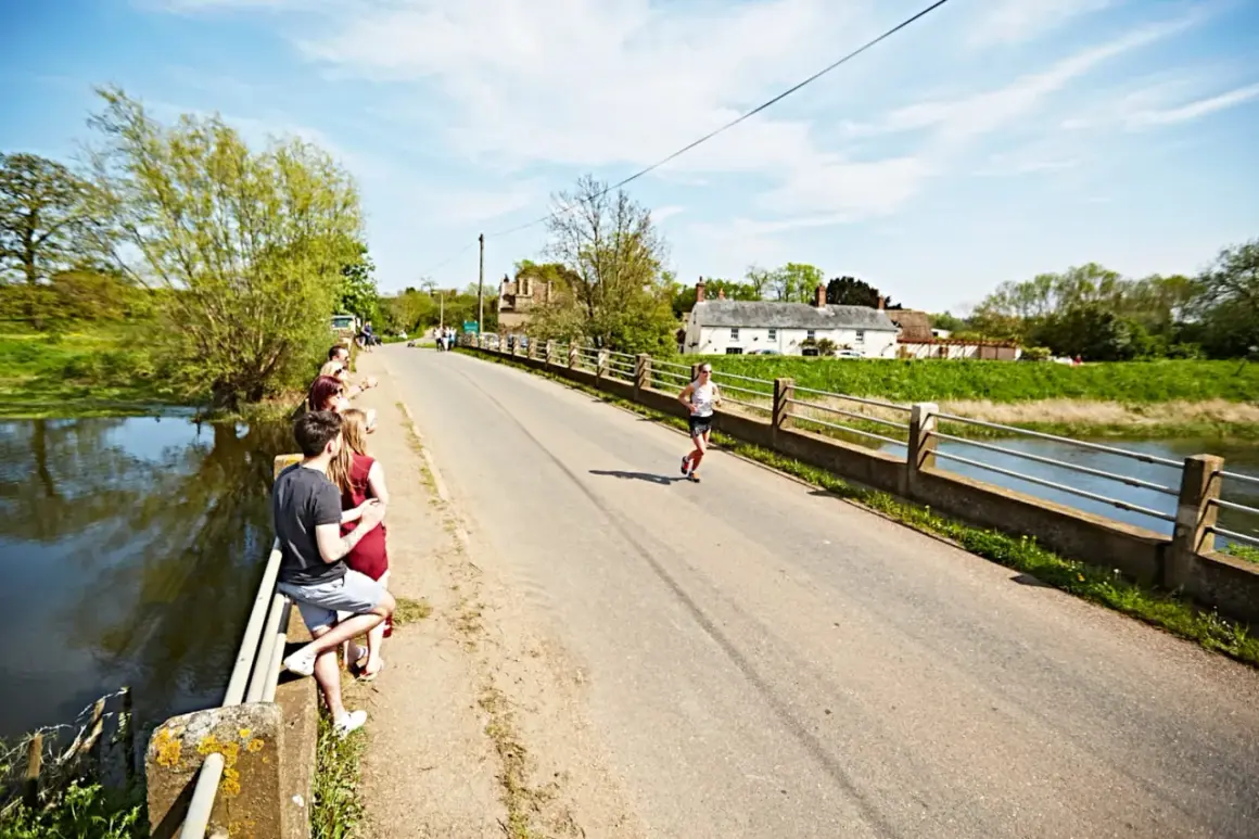 Une femme court sur une route ensoleillée, traversant un pont bordé d'arbres. Des spectateurs encouragent la course à pied en Bretagne.