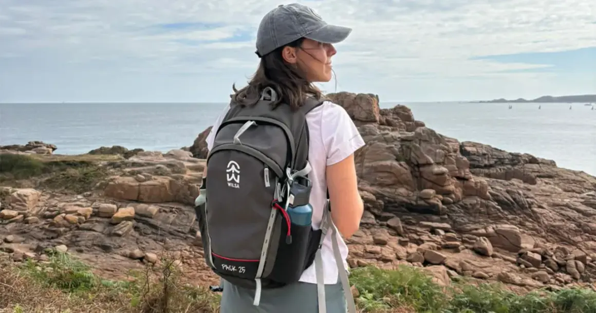Jeune femme avec un sac à dos, prête à faire de la randonnée sur un sentier côtier rocheux, face à la mer.