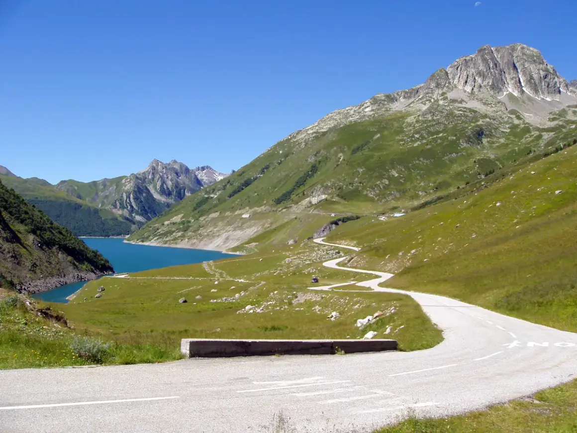 Route sinueuse menant au col du Glandon, avec un lac bleu vif et des montagnes verdoyantes sous un ciel clair.