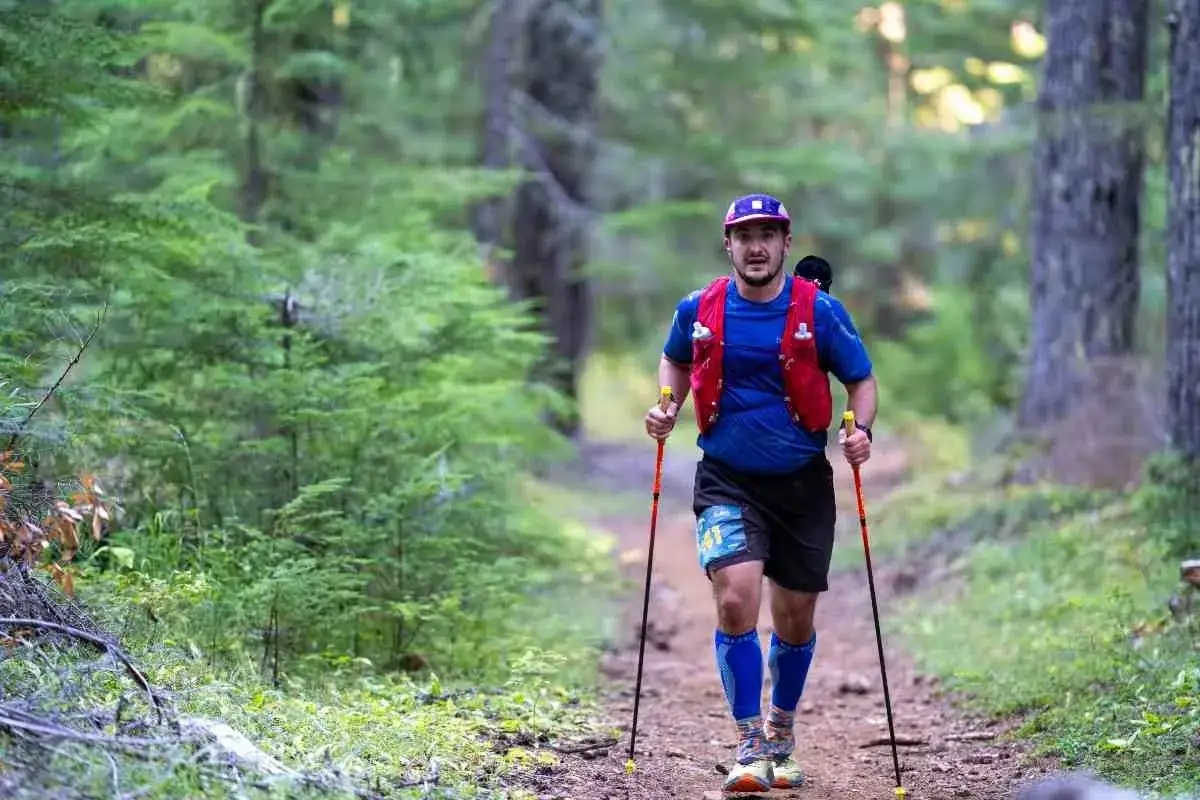 Un homme en tenue de sport, casquette et gilet rouge, pratique le fast hiking en for&ecirc;t, b&acirc;tons &agrave; la main.