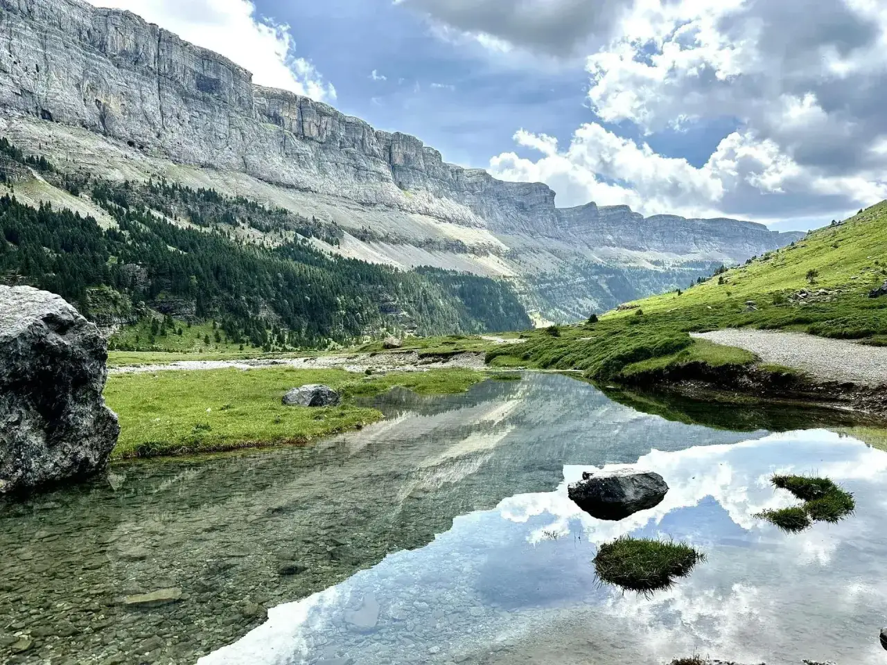 Magnifique paysage de randonnée montagne avec un lac reflétant les falaises et les nuages. Un chemin serpente sur la colline verdoyante.
