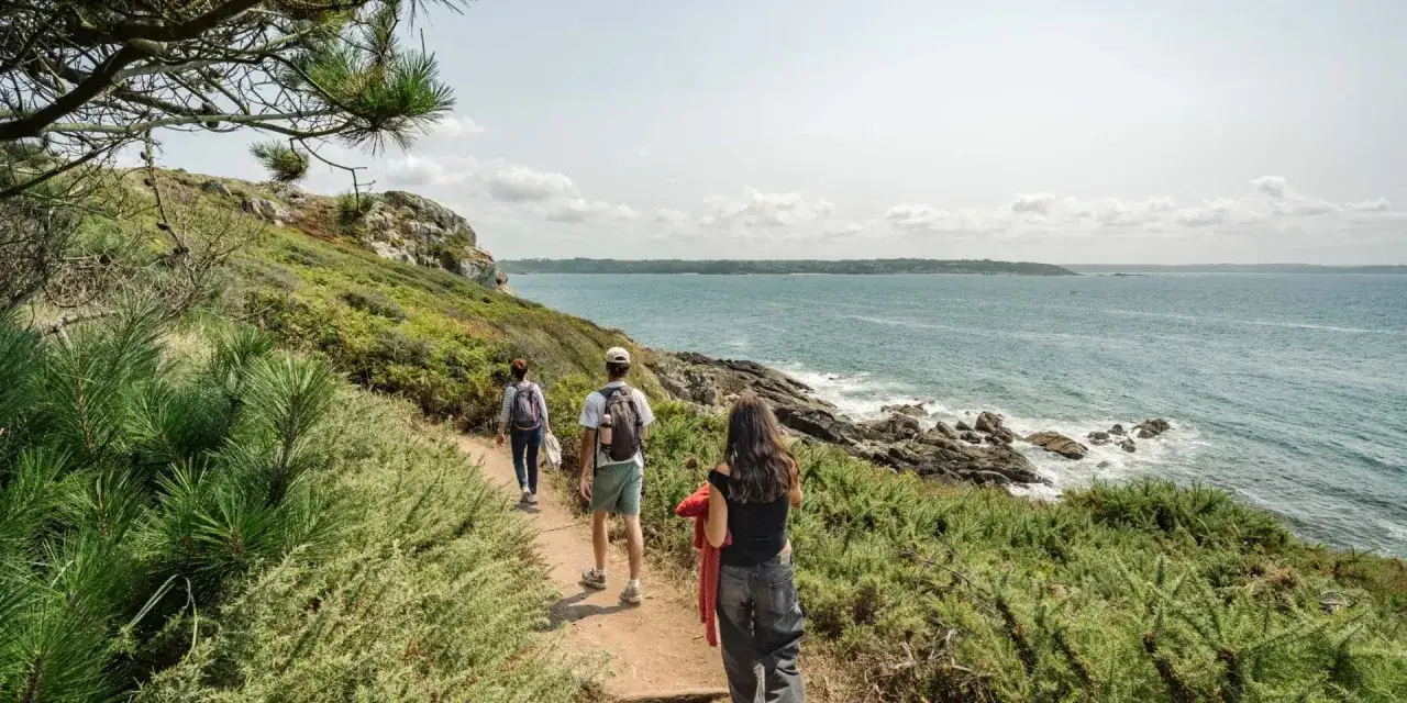 Trois randonneurs suivent le sentier côtier, un record gr34 à la main, longeant la mer d'un bleu profond.