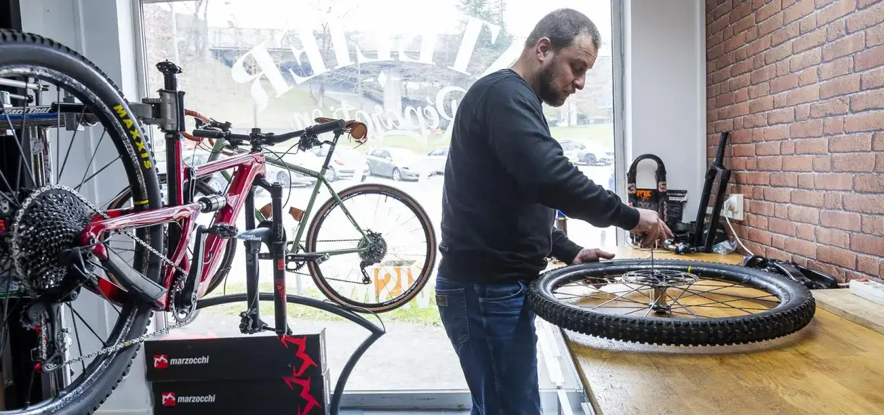 Un m&eacute;canicien effectue un entretien VTT, travaillant sur une roue de v&eacute;lo pos&eacute;e sur un &eacute;tabli. D'autres v&eacute;los sont visibles en arri&egrave;re-plan.