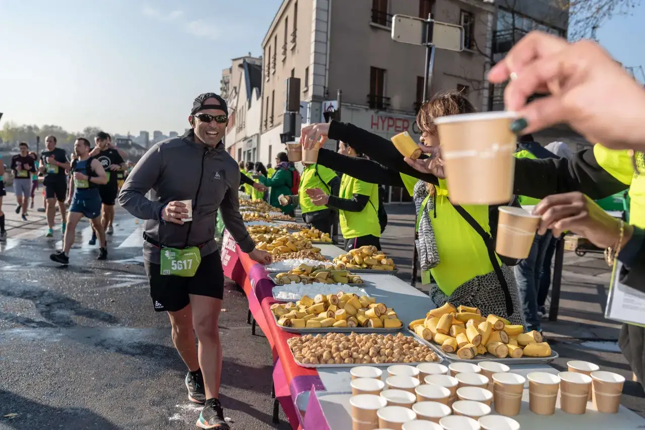 Un coureur souriant prend une coupe lors d'un semi-marathon, près d'une table garnie de bananes et de sucres.