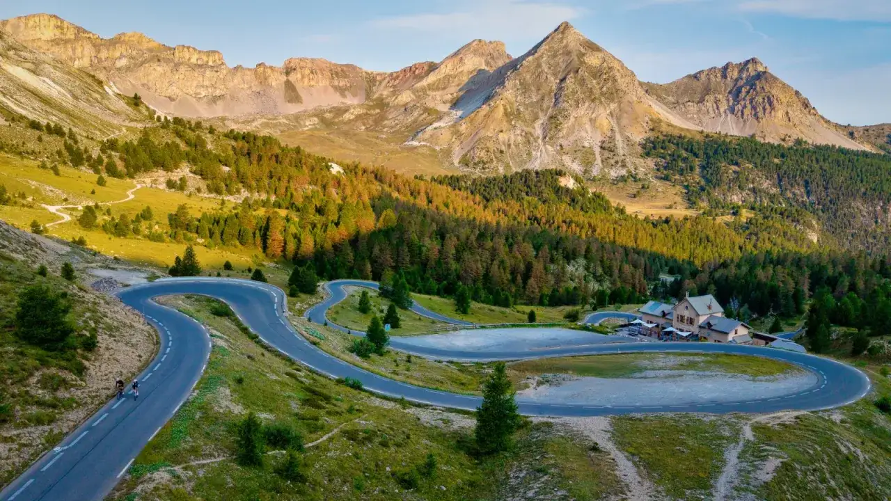 Route sinueuse du col d'Izoard, avec des cyclistes gravissant la pente, des montagnes majestueuses et une for&ecirc;t d'&eacute;pic&eacute;as.