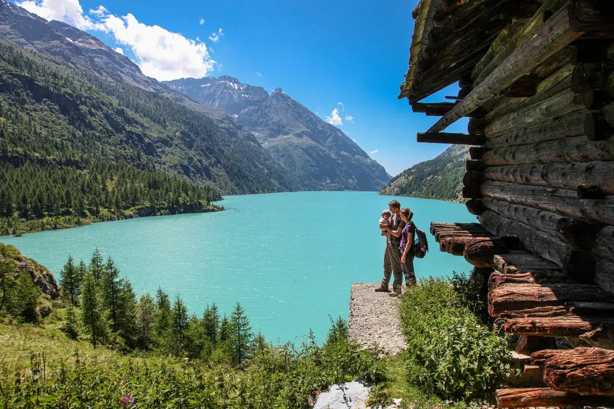 Famille en randonnée à Aoste, admirant un lac turquoise entouré de montagnes et d'une cabane en bois.