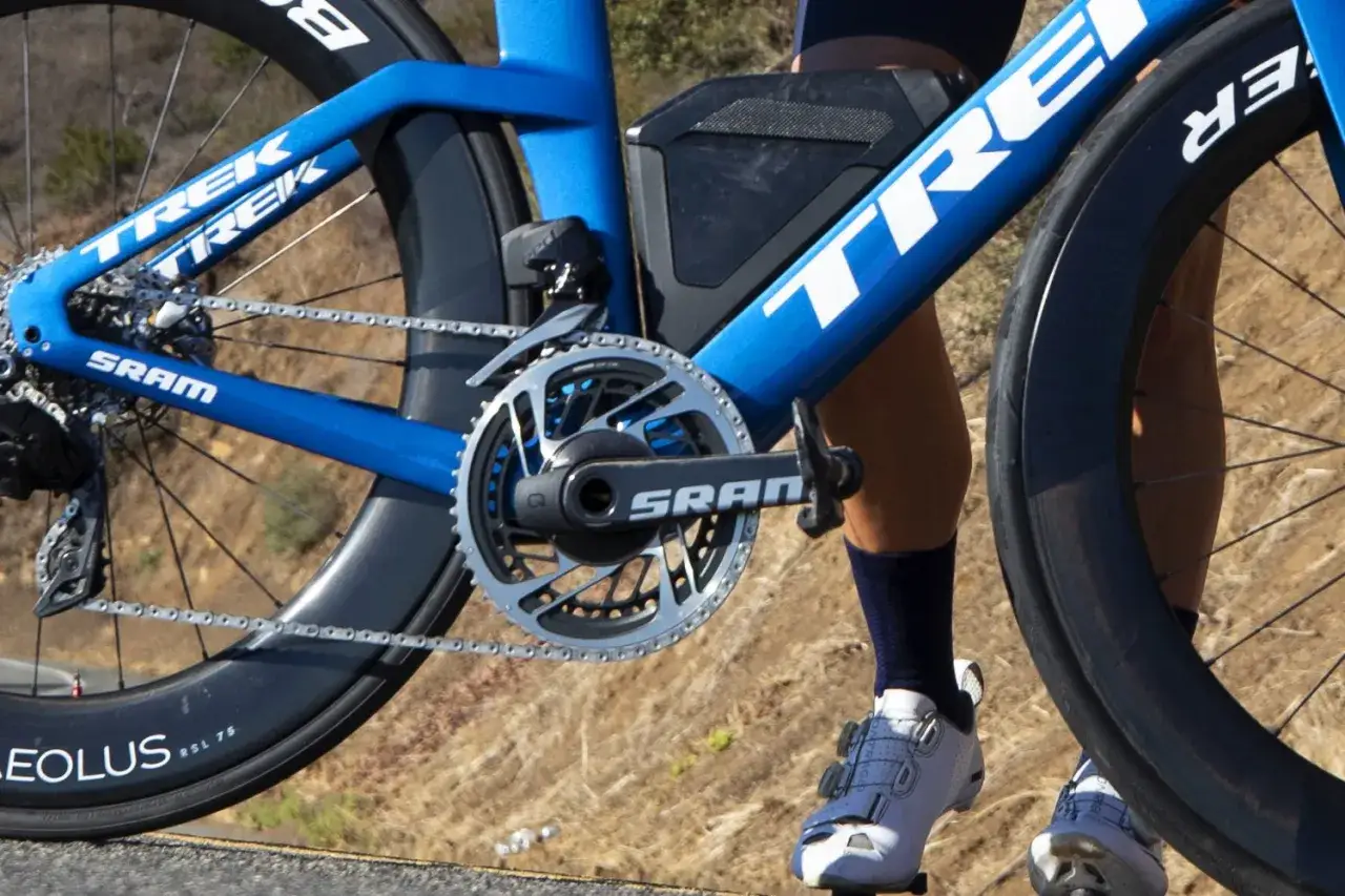 D&eacute;monter p&eacute;dale v&eacute;lo : gros plan sur le p&eacute;dalier SRAM d'un v&eacute;lo de course Trek bleu, avec une jambe de cycliste en arri&egrave;re-plan.
