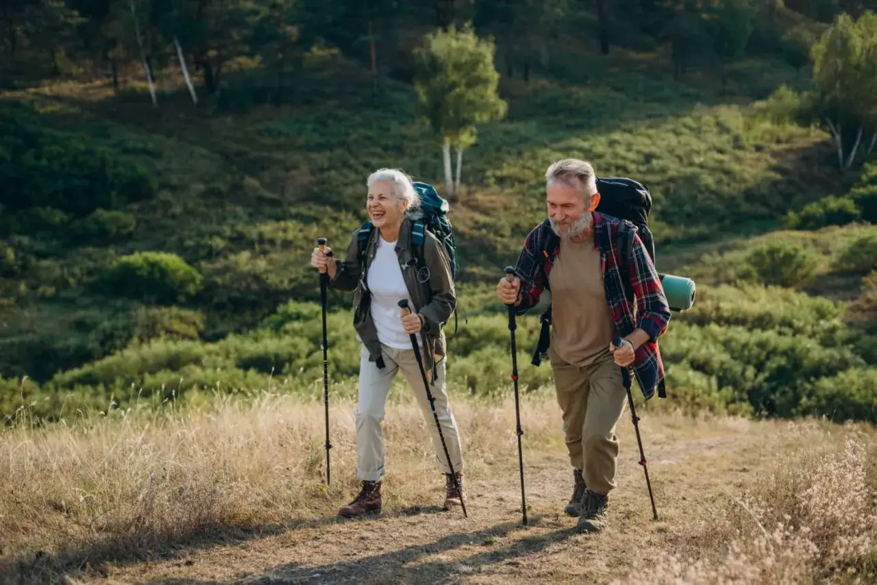 Couple âgé profitant de la marche nordique en pleine nature, souriants et équipés de bâtons et sacs à dos.