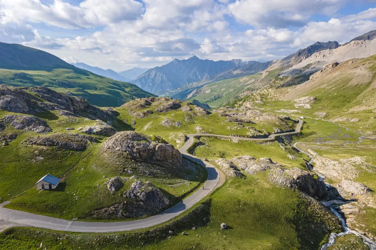 Vue a&eacute;rienne du col de la Bonette, avec sa route sinueuse traversant des paysages alpins verdoyants et rocheux. Un cycliste solitaire grimpe vers le sommet.